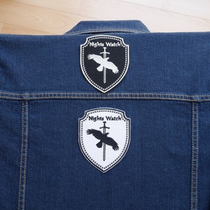 Peut inclure: Une veste en jean bleue avec deux patchs brodés en noir et blanc. Les patchs sont en forme de boucliers et présentent un corbeau avec une épée et le texte "Nights Watch".