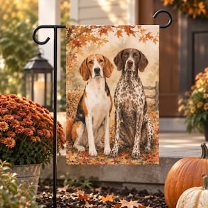 Könnte beinhalten: Eine rechteckige Gartenflagge mit einer Aquarellillustration von zwei Hunden in einer herbstlichen Landschaft. Ein Hund ist ein dreifarbiger Laufhund mit lohfarbenem, schwarzem und weißem Fell, der andere ein braun-weiß gefleckter Vorstehhund.