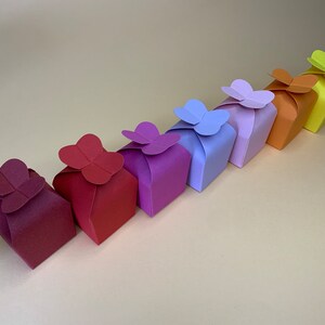 Könnte beinhalten: Eine Reihe farbenfroher Geschenkboxen in den Farbtönen Burgunderrot, Rot, Lila, Lavendel, Orange und Gelb. Jede Box ist mit einem Papier-Schmetterling verziert. Die Boxen sind diagonal auf einer hellbeigen Oberfläche angeordnet.