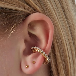 May include: A gold ear cuff, adorned with a continuous line of small, clear gemstones. Two small, gold stud earrings are also visible on the ear. The ear is set against a backdrop of blonde hair.