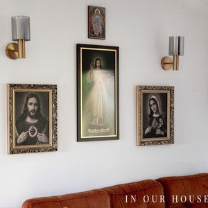 May include: A white wall with four framed religious images and two gold wall sconces with glass shades. The images depict Jesus, Mary, and other religious figures. The text "IN OUR HOUSE:" is visible at the bottom of the image.