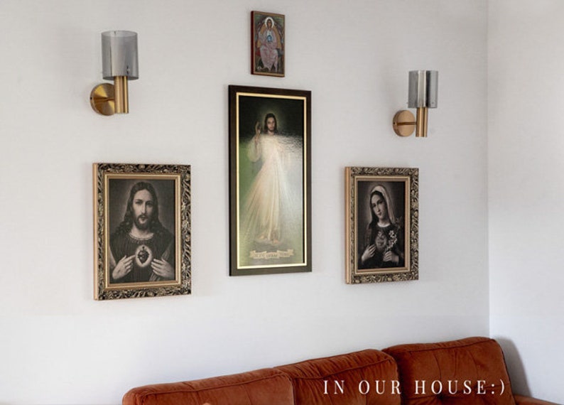 May include: A white wall with four framed religious images and two gold wall sconces with glass shades. The images depict Jesus, Mary, and other religious figures. The text "IN OUR HOUSE:" is visible at the bottom of the image.
