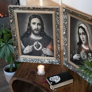 May include: Two framed religious images, one depicting Jesus Christ and the other depicting the Virgin Mary, both with ornate silver frames. A lit candle and a rosary are placed on a wooden table in front of the images.