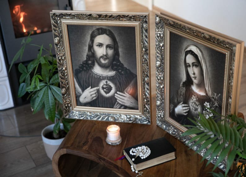 May include: Two framed religious images, one depicting Jesus Christ and the other depicting the Virgin Mary, both with ornate silver frames. A lit candle and a rosary are placed on a wooden table in front of the images.
