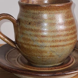 May include: A handcrafted ceramic mug and saucer. The mug, with a curved handle, is a warm brown with horizontal bands. The saucer is a matching brown with a slightly raised edge. The set is on a wooden surface.