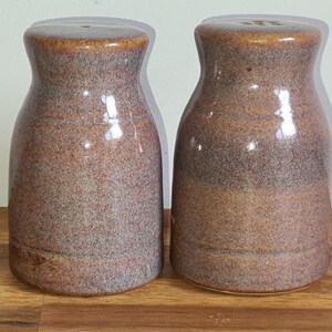 May include: Two ceramic salt and pepper shakers. The shakers are brown with a speckled texture and a glossy finish. The tops are a darker brown. They are sitting on a wooden surface.