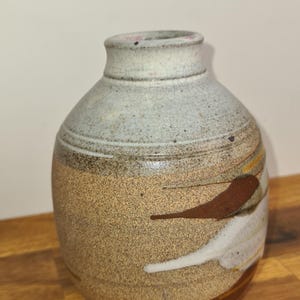 May include: A ceramic vase with a rounded body and a narrow neck. The vase has a speckled tan and brown exterior with abstract brown and white painted accents. The top is a light grey colour.