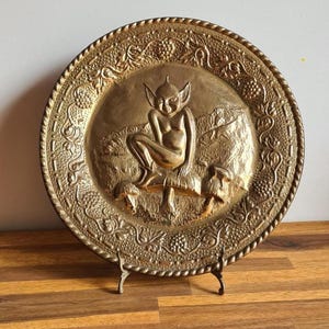 May include: A vintage brass decorative plate with a relief of a seated fairy figure. The plate has a detailed border with floral and grape vine designs. The plate is displayed on a small metal stand, showcasing its intricate craftsmanship and antique aesthetic.