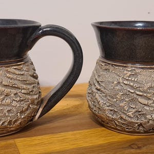May include: Two handmade ceramic mugs. The top and handle are dark grey, while the lower portion features a textured, bark-like design in a light tan colour. The mugs are displayed on a wooden surface.