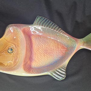 May include: A ceramic fish-shaped serving dish with a glazed finish. The dish is decorated with a realistic fish design in shades of pink, orange, and green.