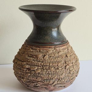 May include: A ceramic vase with a textured, bark-like exterior in shades of brown and tan. The vase has a dark green, glossy neck and rim, and a bulbous body. A decorative piece for home decor.