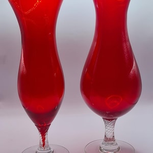 May include: Two vivid red glass vases. Each vase features a flared top, a rounded body, and a clear, twisted stem leading to a circular base. The vases are a striking shade of red and appear to be made of glass.