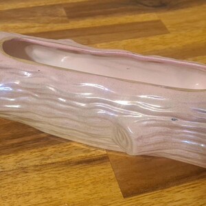 May include: A light pink ceramic planter in the shape of a log. The planter features a rectangular opening and a textured surface designed to resemble tree bark. The interior is a lighter shade of pink.