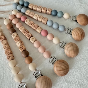 May include: A collection of personalized baby pacifier clips. Each clip features wooden beads in various colors, including beige, pink, and blue, with wooden letter beads spelling out names. The clips have a wooden round clip and a metal clasp.