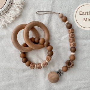 May include: Wooden baby teething toys and pacifier clip in an earthy color palette. The set includes two wooden rings, a beaded teether with the name "EMMA", and a pacifier clip with a sunflower detail. The image has a neutral background.