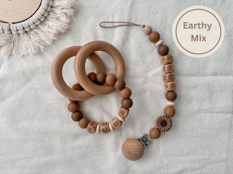May include: Wooden baby teething toys and pacifier clip in an earthy color palette. The set includes two wooden rings, a beaded teether with the name "EMMA", and a pacifier clip with a sunflower detail. The image has a neutral background.