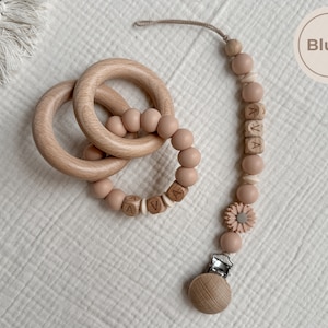 May include: A wooden baby teether set in blush pink. The set includes two wooden rings, a beaded teether with the name "AVA" spelled out, and a pacifier clip. The pacifier clip has a wooden clip and a flower accent.