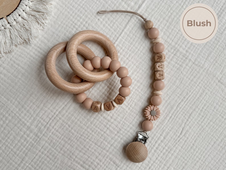 May include: A wooden baby teether set in blush pink. The set includes two wooden rings, a beaded teether with the name "AVA" spelled out, and a pacifier clip. The pacifier clip has a wooden clip and a flower accent.