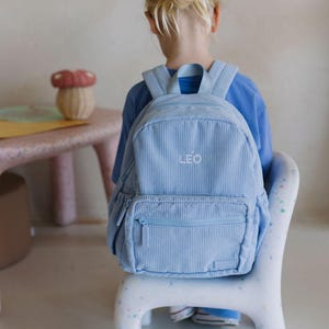 May include: A pale blue corduroy backpack with the name "LEO" embroidered on the front. The backpack features a front zipped pocket and adjustable straps. The child is wearing a blue top and sitting on a white chair.