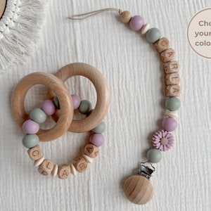 May include: Wooden baby teether and pacifier clip set. The teether features two wooden rings with pastel-colored beads. The pacifier clip has a wooden clip, beads, and a flower accent. The name "CARLA" is spelled out in wooden letter blocks.
