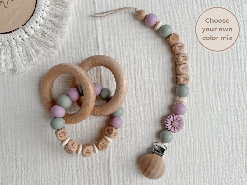 May include: Wooden baby teether and pacifier clip set. The teether features two wooden rings with pastel-colored beads. The pacifier clip has a wooden clip, beads, and a flower accent. The name "CARLA" is spelled out in wooden letter blocks.