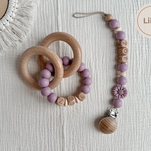 May include: A lilac-colored baby teether set featuring two wooden rings and a pacifier clip. The clip has the name "ZOE" spelled out in wooden beads, along with a flower and a wooden clip. The set is designed for infants.