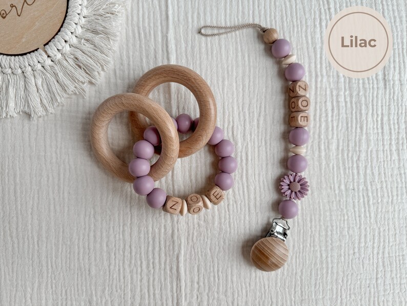 May include: A lilac-colored baby teether set featuring two wooden rings and a pacifier clip. The clip has the name "ZOE" spelled out in wooden beads, along with a flower and a wooden clip. The set is designed for infants.