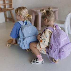 K&ouml;nnte beinhalten: Zwei Kinder mit Rucks&auml;cken, einer hellblau und einer lavendelfarben, sitzen auf einem Skateboard. Auf dem hellblauen Rucksack ist der Name "LEO" gestickt. Auf dem lavendelfarbenen Rucksack steht "HAPPY DAYS".
