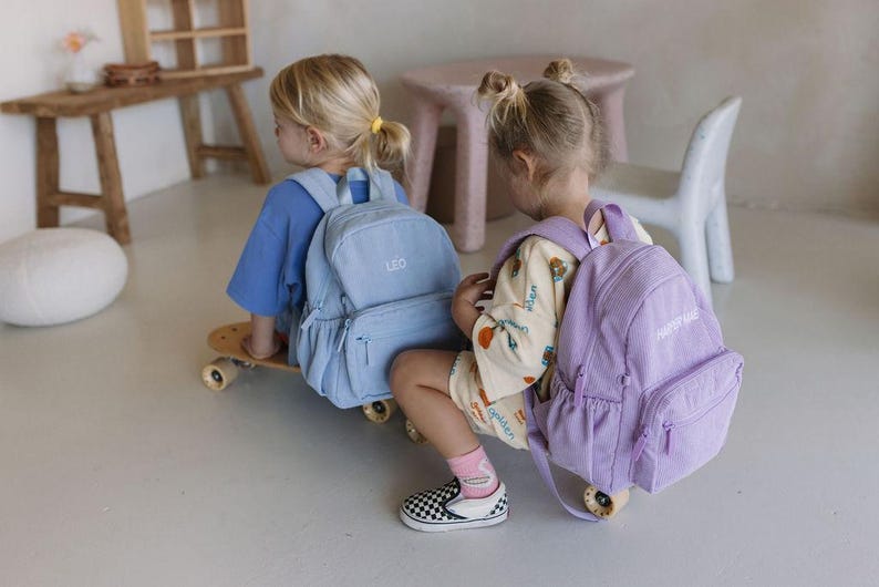 K&ouml;nnte beinhalten: Zwei Kinder mit Rucks&auml;cken, einer hellblau und einer lavendelfarben, sitzen auf einem Skateboard. Auf dem hellblauen Rucksack ist der Name "LEO" gestickt. Auf dem lavendelfarbenen Rucksack steht "HAPPY DAYS".