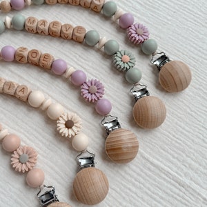 May include: Several baby pacifier clips with wooden beads and floral accents. Each clip features a wooden clip, a string of beads in pastel colors, and a name spelled out in wooden letter blocks. The clips have a daisy flower design.