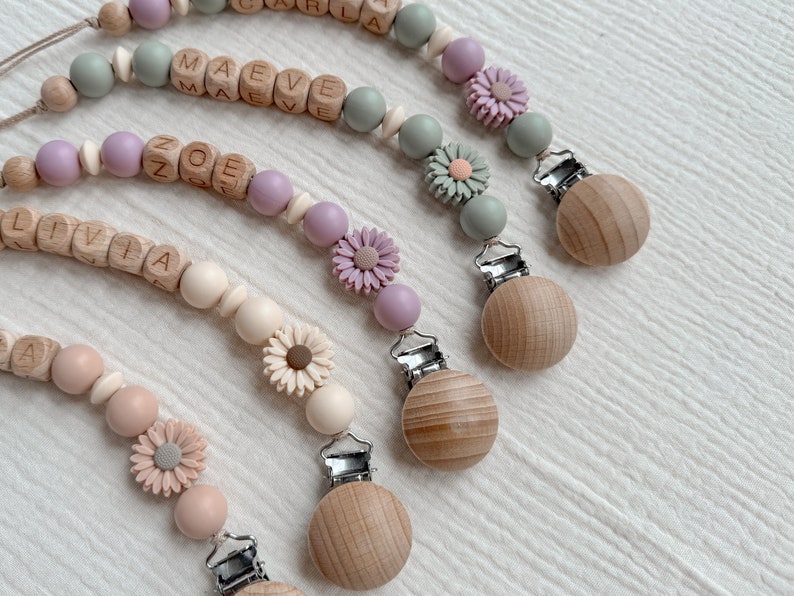 May include: Several baby pacifier clips with wooden beads and floral accents. Each clip features a wooden clip, a string of beads in pastel colors, and a name spelled out in wooden letter blocks. The clips have a daisy flower design.