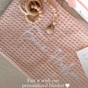 May include: A pink waffle-weave baby blanket with the name "Emma Hudson" embroidered in white. A wooden teething ring and pacifier clip with the name "Emma" are on the blanket. The text "Pair it with our personalized blanket" is also visible.