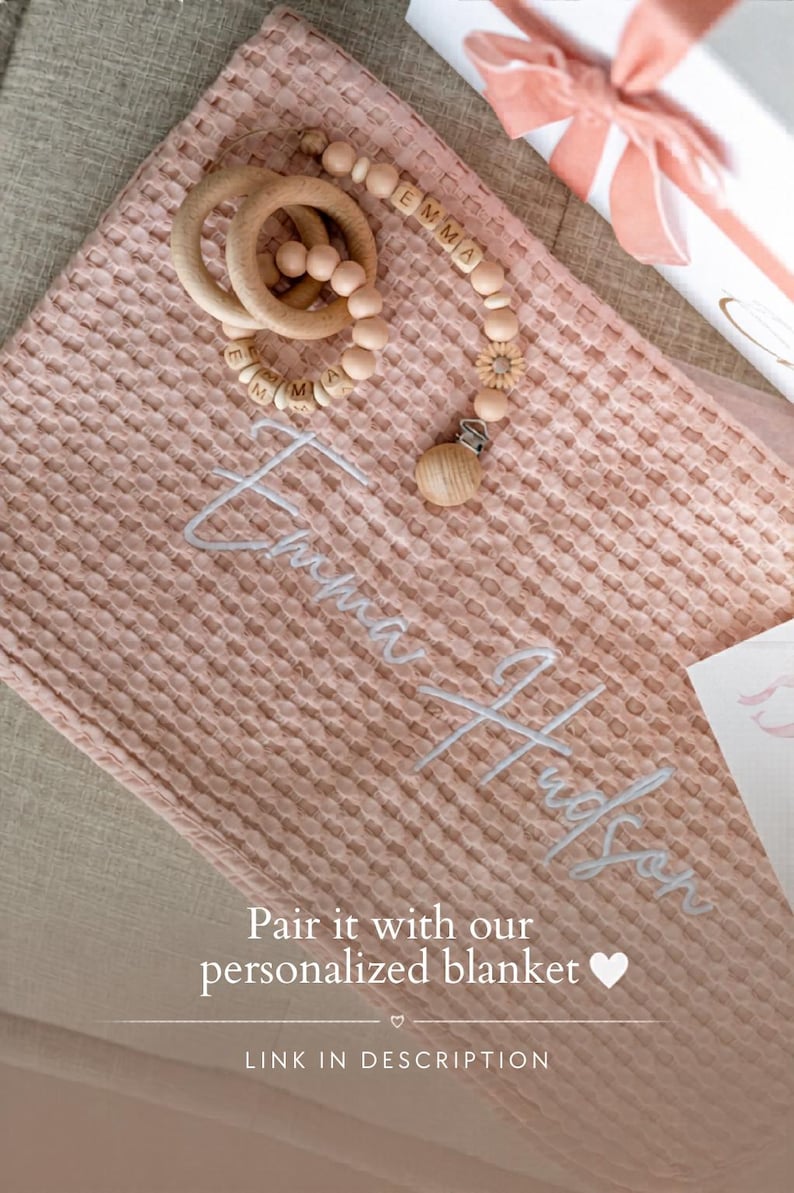 May include: A pink waffle-weave baby blanket with the name "Emma Hudson" embroidered in white. A wooden teething ring and pacifier clip with the name "Emma" are on the blanket. The text "Pair it with our personalized blanket" is also visible.