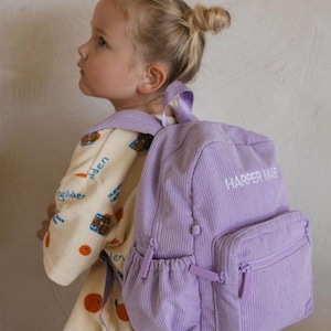 May include: A lilac corduroy backpack with the text "HARPER MAE" embroidered in white. The backpack features a front pocket and side pocket with zips. The bag has a top handle and adjustable shoulder straps.