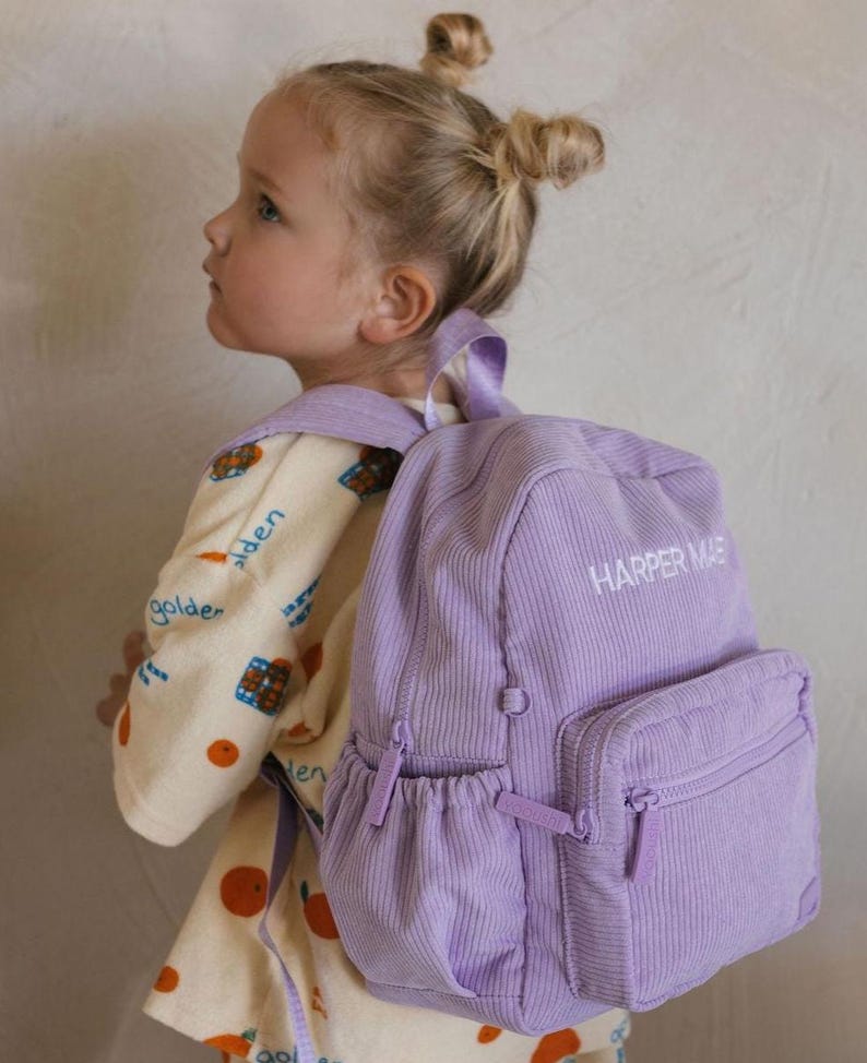 K&ouml;nnte beinhalten: Ein lavendelfarbener Cord-Rucksack mit dem Text "HARPER MAE" in Wei&szlig; gestickt. Der Rucksack hat eine Vordertasche und eine Seitentasche mit Rei&szlig;verschl&uuml;ssen. Die Tasche hat einen oberen Griff und verstellbare Schultergurte.