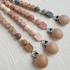 May include: Four personalized wooden baby teethers with silicone beads and clips. The teethers are made with natural wood beads and feature the names Jaxson, Wyatt, Emma, and Noah. Each teether has a silicone rainbow charm and a wooden clip.