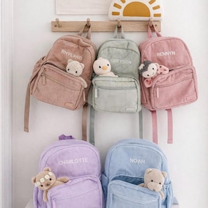 May include: A collection of corduroy backpacks in various pastel colors, each personalized with a name. The backpacks are hanging on a wooden rack, with stuffed animals peeking out of the front pockets. A sign that says "SUNSHINE" is above the backpacks.