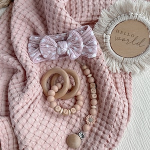 May include: A baby gift set featuring a pink waffle-knit blanket, a polka-dot headband, wooden teething rings, and a pacifier clip with the name "AVA". A wooden disc with the words "HELLO world" is also included.