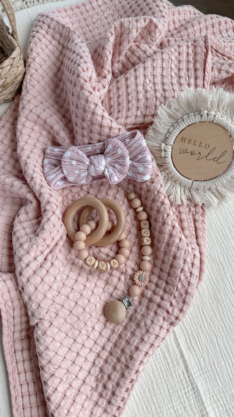 May include: A baby gift set featuring a pink waffle-knit blanket, a polka-dot headband, wooden teething rings, and a pacifier clip with the name "AVA". A wooden disc with the words "HELLO world" is also included.