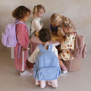 K&ouml;nnte beinhalten: Kinder mit Rucks&auml;cken aus Cordstoff in verschiedenen Pastellfarben. Die Rucks&auml;cke haben eine Vordertasche und verstellbare Tr&auml;ger. Auf einem Rucksack steht der Name "LEO". Die Kinder sitzen um einen kleinen Tisch.