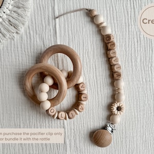 May include: A cream-colored baby pacifier clip and wooden rattle set. The pacifier clip features wooden beads and the name "OLIVIA" spelled out in wooden letter beads. The rattle has wooden rings and beads.