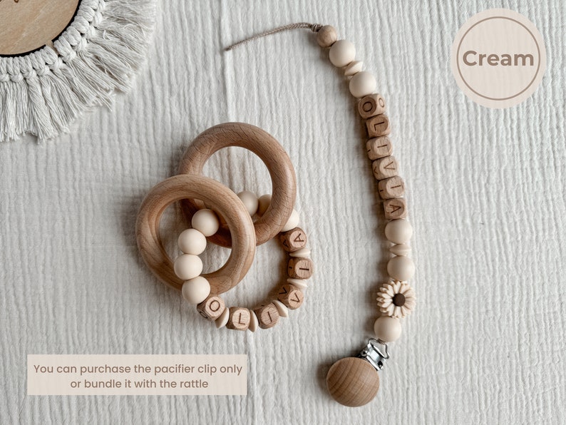 May include: A cream-colored baby pacifier clip and wooden rattle set. The pacifier clip features wooden beads and the name "OLIVIA" spelled out in wooden letter beads. The rattle has wooden rings and beads.