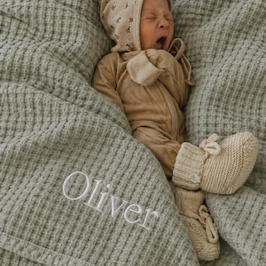 May include: A newborn baby wearing a beige onesie and bonnet with a floral pattern, lying on a sage green waffle-weave blanket. The blanket is embroidered with the name "Oliver" in white. The baby is also wearing cream-colored knitted booties.