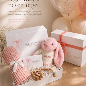 May include: A gift box with a pink bunny, a pink blanket, and a wooden teether. The box includes a card that says "WELCOME". The text "Emma" is on the bunny and the box. The box is white with a pink ribbon.