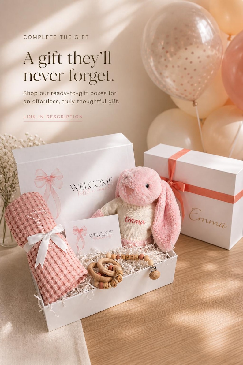 May include: A gift box with a pink bunny, a pink blanket, and a wooden teether. The box includes a card that says "WELCOME". The text "Emma" is on the bunny and the box. The box is white with a pink ribbon.