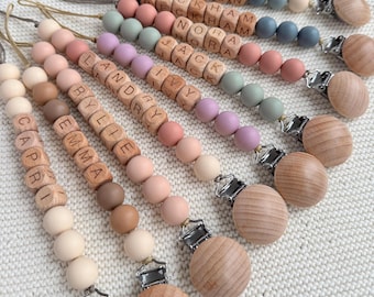 Pode incluir: Um conjunto de dez mordedores de bebê de madeira personalizados com contas de silicone. Cada mordedor apresenta um nome diferente escrito em contas de madeira e um clipe de madeira. Os mordedores são de várias cores, incluindo rosa, azul, verde e marrom.