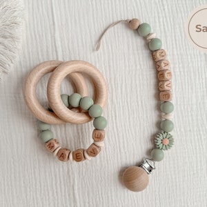 May include: A wooden baby teether and pacifier clip set in sage green. The teether features two wooden rings and sage green beads. The pacifier clip has wooden beads spelling "MAEVE" and a daisy accent.