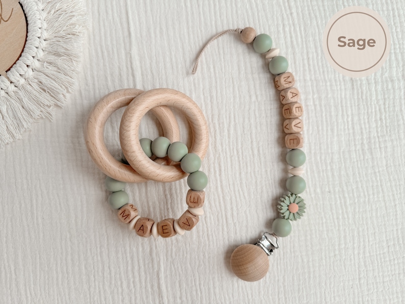 May include: A wooden baby teether and pacifier clip set in sage green. The teether features two wooden rings and sage green beads. The pacifier clip has wooden beads spelling "MAEVE" and a daisy accent.