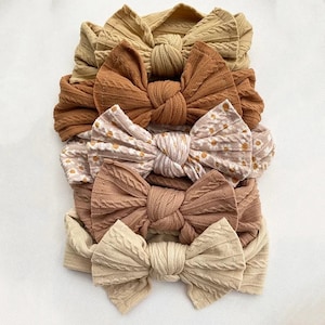 May include: A stack of six baby headbands in various shades of brown and beige. The headbands feature a knotted bow design, with some having a textured cable knit pattern and others a floral print. The headbands are stacked on a white surface.