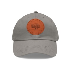 Könnte beinhalten: Graue Baseballkappe mit einem braunen Lederaufnäher mit einer Baumillustration und dem Text "THE TREE GIVETH, THE TREE TAKETH AWAY". Die Kappe hat einen gebogenen Schirm und einen Stoffriemenverschluss.